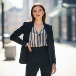 Mladá žena v elegantním oblečení stojí ve městě, jemné světlo zdůrazňuje její postavu.